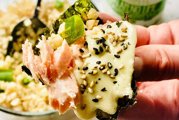 Hand holding a nori sheet filled with flaked cooked salmon, chopped green onions, cauliflower rice, Primal Kitchen Jalapeno Lime Mayo with black and white sesame seeds