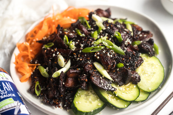 Crispy pork belly pieces in Primal Kitchen Korean Style BBQ Sauce with carrot ribbons and cucumbers, on a white plate. 