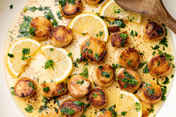 Seared scallops in a white skillet with avocado oil, lemon slices, chopped parsley and a wooden spatula