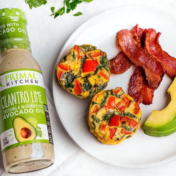 Two veggie egg bites, bacon strips, and sliced avocado on a plate next to a bottle of Primal Kitchen Cilantro Lime Dressing. 