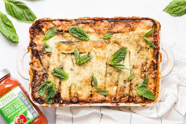 Overhead shot of zucchini lasagna with fresh basil leaves, beside a jar of Primal Kitchen Tomato Basil Marinara. 