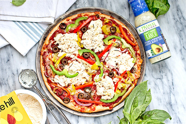 Vegan pizza with olives, peppers, and Kite Hill Ricotta next to a bottle of Primal Kitchen Plant Based Ranch.