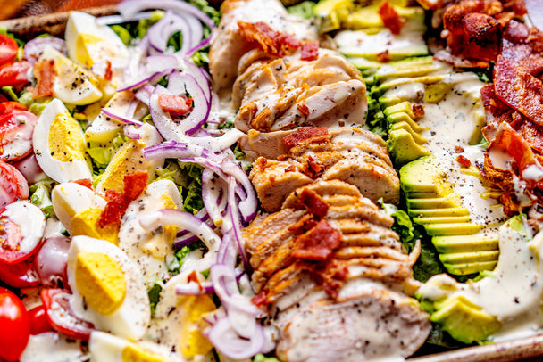 Very close up shot of sheet pan cobb salad made with romaine lettuce, Primal Kitchen Ranch Dressing, red onions, sliced chicken, sliced avocado, chopped cherry tomatoes, quartered hard boiled eggs, and crumbled bacon