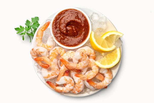 Overhead view of a clear plate with chilled shrimps and a bowl of Primal Kitchen cocktail sauce.