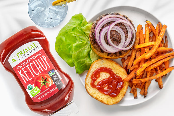 A burger open on a plate with sweet potato fries, next to a bottle of Primal Kitchen Squeeze Ketchup.