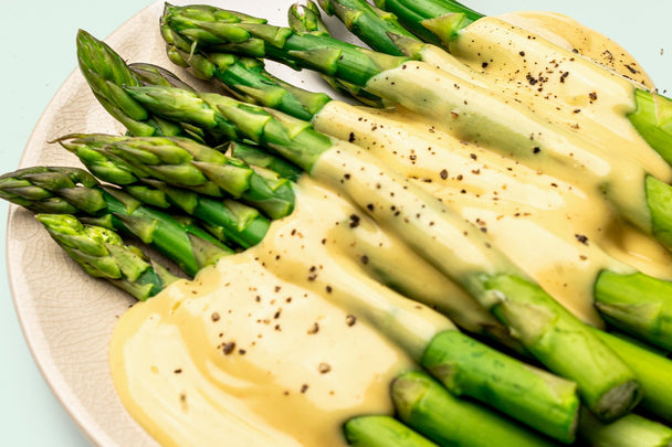 Close up shot of a white plate of steamed asparagus topped with Hollandaise Sauce and cracked black pepper.