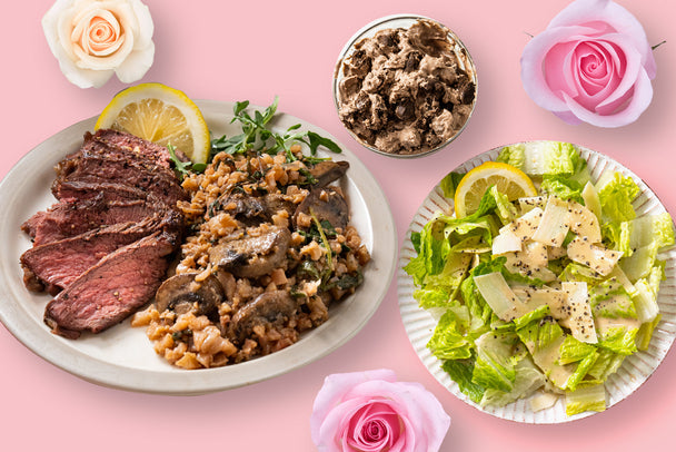 Valentine's Day dinner at home three-course meal: Sliced steak and cauliflower risotto with mushrooms on a white plate, Caesar salad on a white plate, and chocolate cookie dough ice cream in a white bowl. White and pink roses in the background.