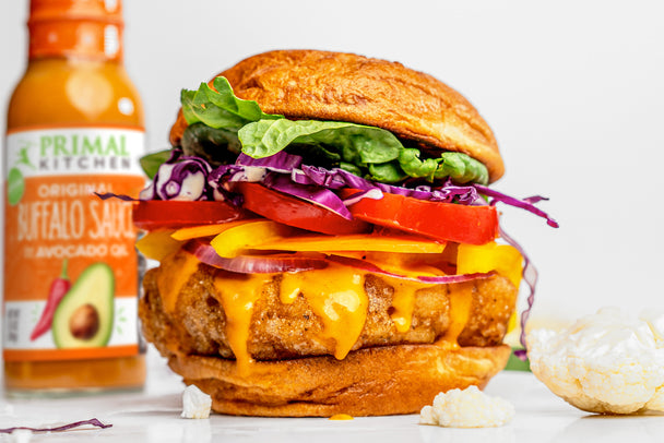 A vegan buffalo cauliflower sandwich drips with buffalo sauce and sits next to a buffalo sauce bottle and cauliflower florets.