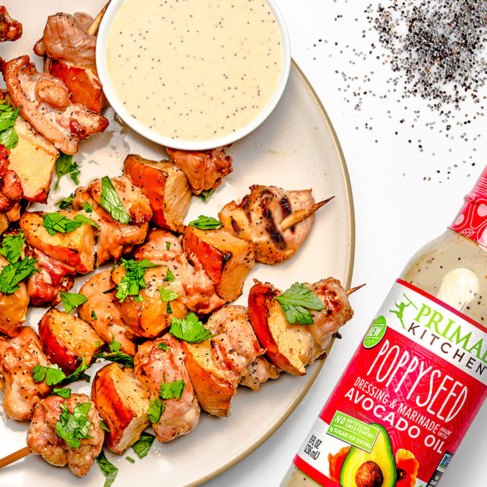 Grilled chicken apple skewers and a bowl of dressing on a white plate, next to a bottle of Primal Kitchen Poppyseed Dressing.