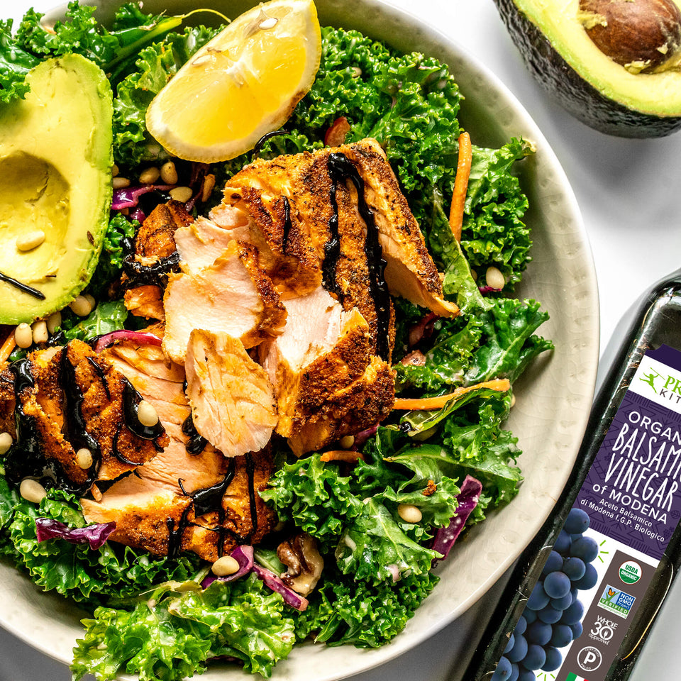 Cut salmon and kale salad with a lemon and avocado in a white bowl next to a cut avocado and bottle of Primal Kitchen Organic Balsamic Vinegar of Modena.