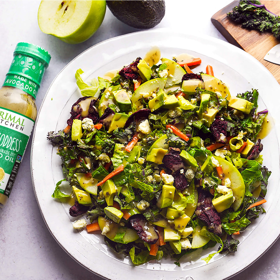 A delicious beet salad with avocado and green apples on a white plate next to a bottle of Primal Kitchen Green Goddess Dressing made with avocado oil.  