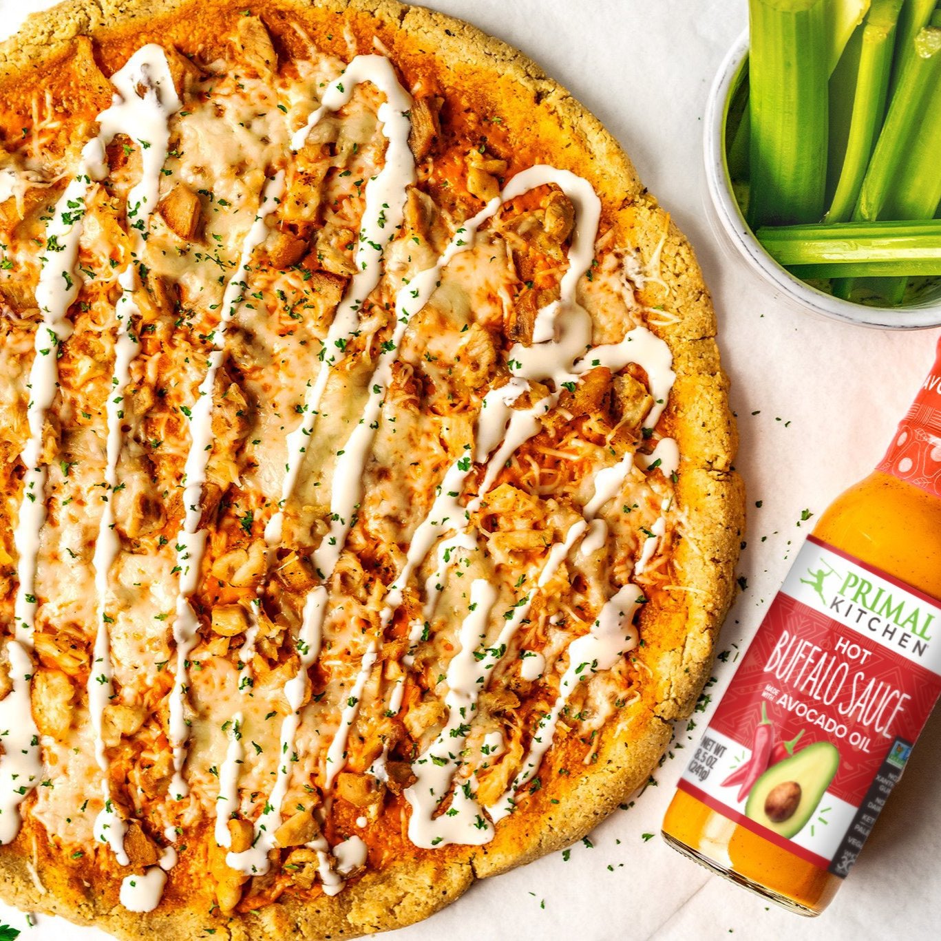 Hot Buffalo Pizza drizzled with ranch dressing, alongside Primal Kitchen Hot Buffalo Sauce and cup of chopped celery