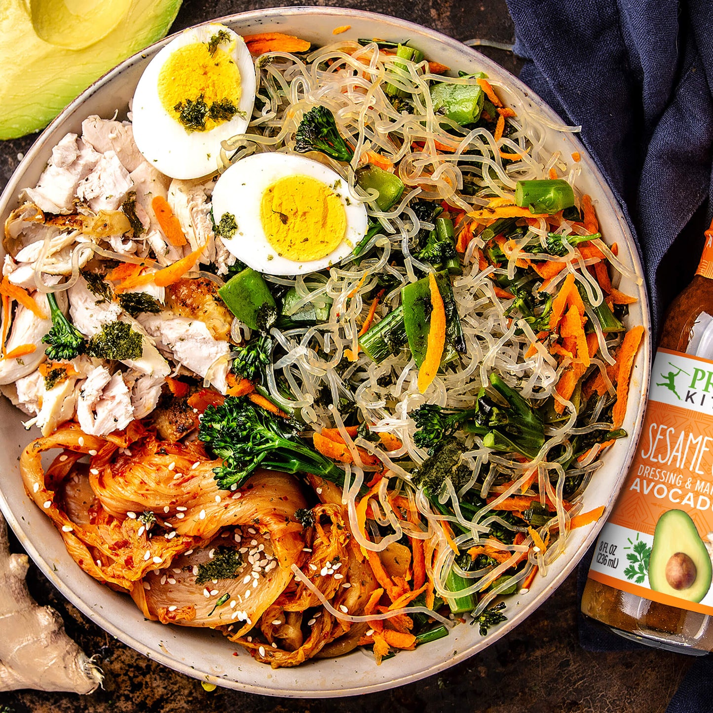 An Asian chicken noodle bowl with egg and avocado , next to a bottle of Sesame Ginger vinaigrette dressing. 