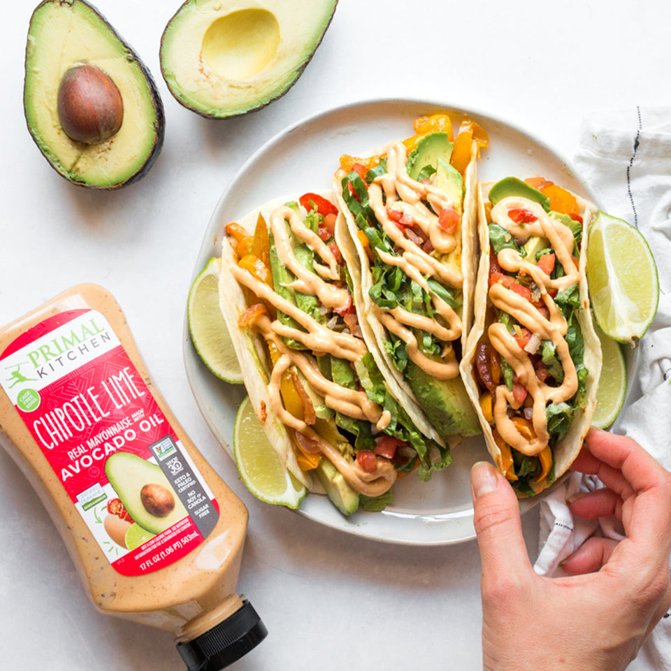 Three veggie flour tacos drizzled with chipotle lime mayo on a white plate with cut up limes next to a cut avocado and a squeeze bottle of Primal Kitchen Chipotle Lime Mayo.